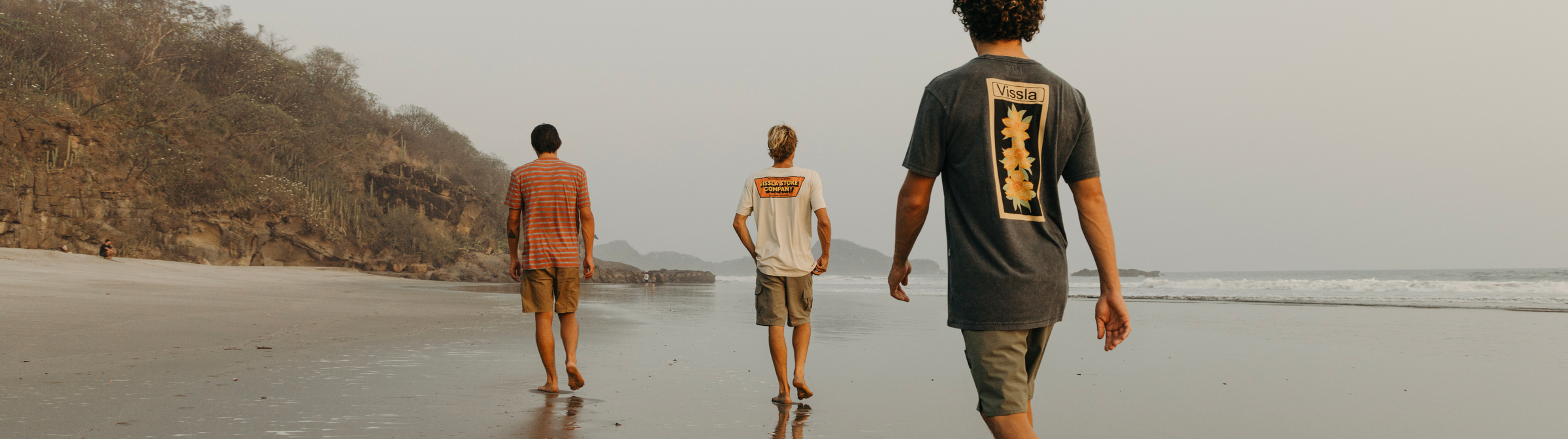 A surfer sitting on a curb in a Mexican village wearing a Vissla tee shirt and hybrid walkshorts